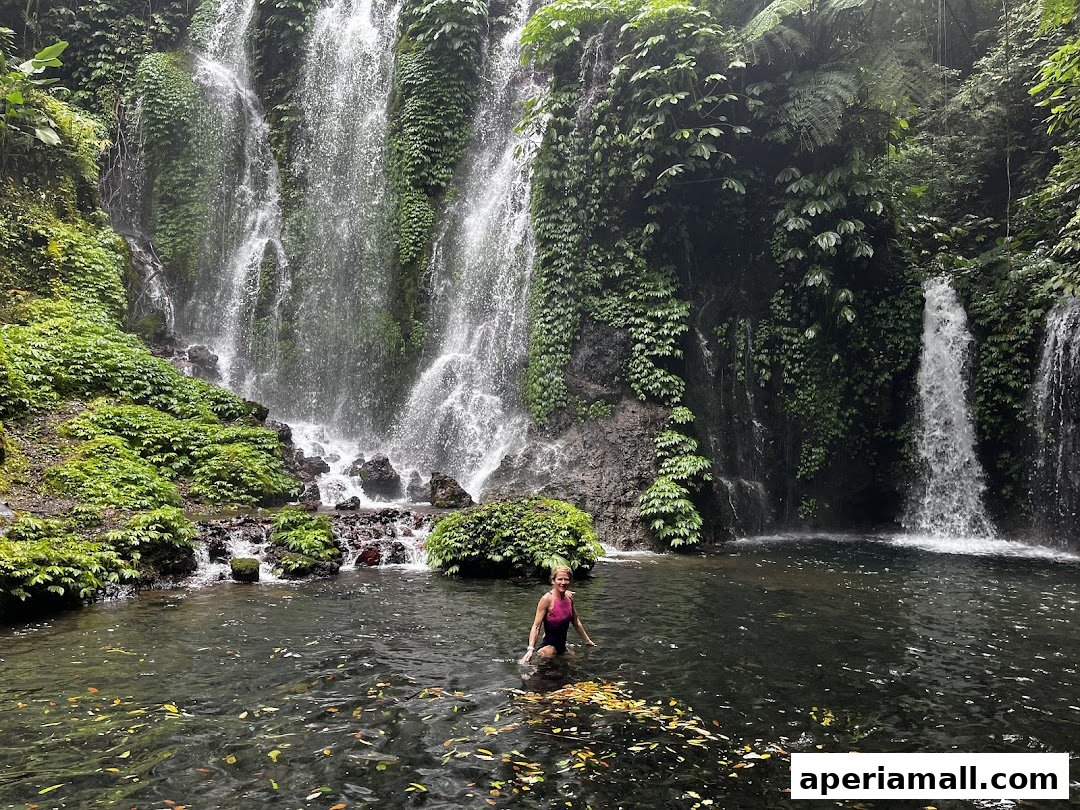 A woman stands in a natural pool at the base of a lush, multi-tiered waterfall.