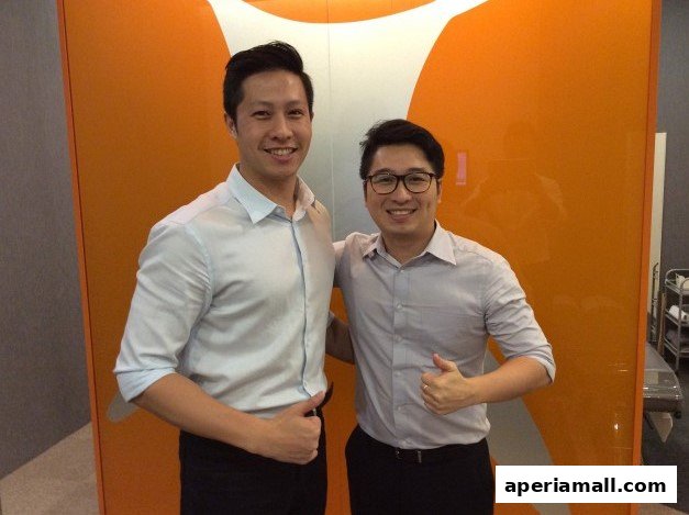 Two men in light-colored shirts and dark pants give thumbs up in front of an orange wall with a logo.