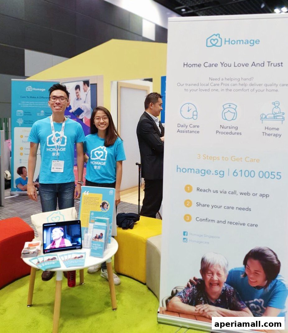 Two smiling staff members in blue t-shirts stand next to a display for Homage Home Care.
