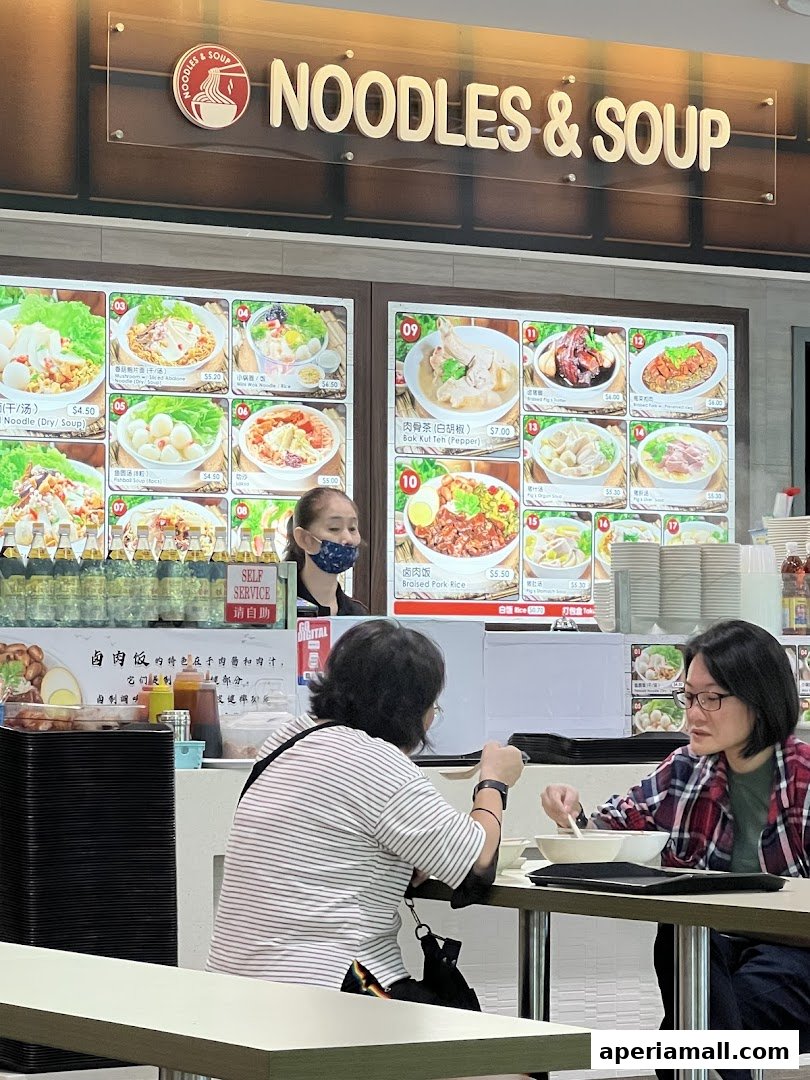 People dining at a food stall with a menu board displaying various noodle and soup dishes.