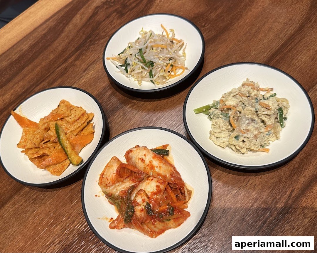 Four small bowls of Korean side dishes, including kimchi, bean sprouts, and fish cakes, are arranged on a wooden table.