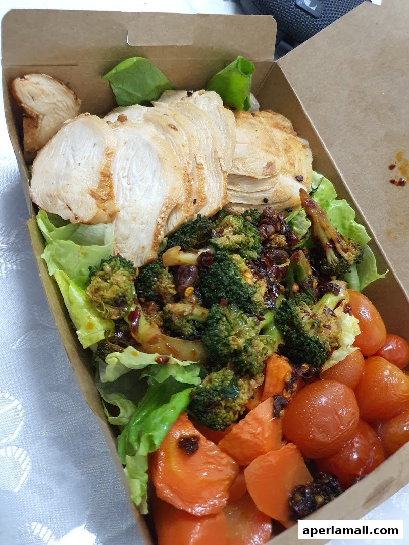 A healthy salad in a takeaway box with chicken, broccoli, carrots, and tomatoes.