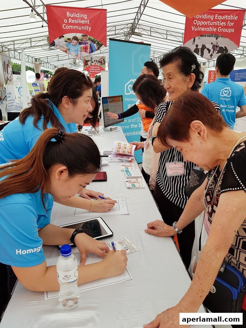 Staff assist visitors at a Homage Home Care booth, with banners about community and opportunities.