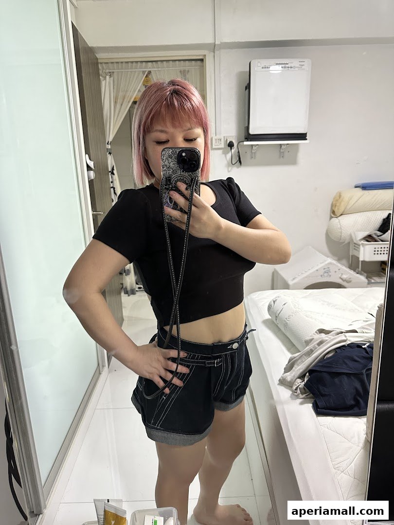 A person with pink hair takes a mirror selfie wearing a black crop top and denim shorts.