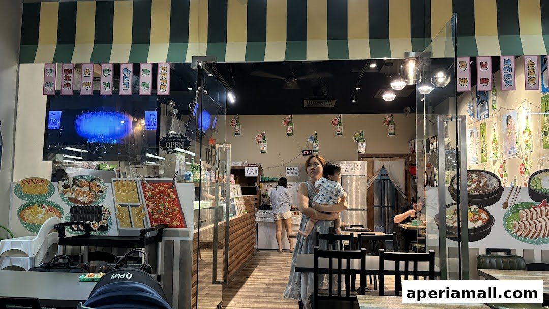 A woman holds a baby in front of a Korean restaurant with food displays and seating.