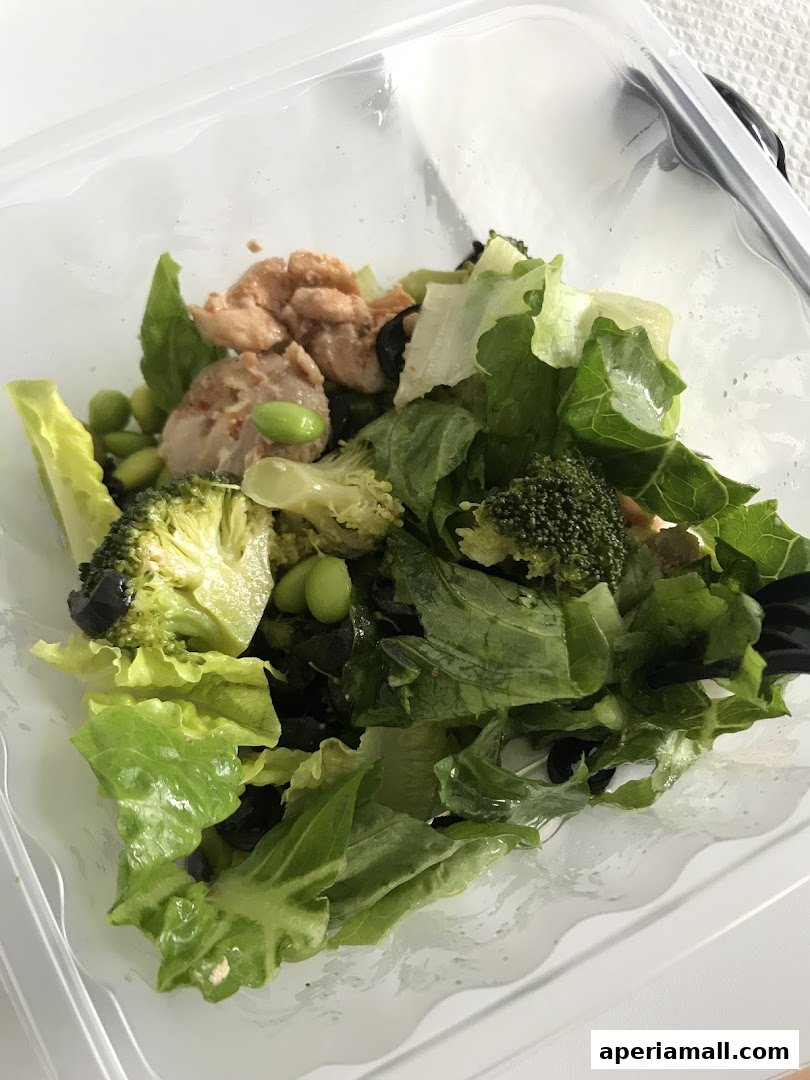 A close-up of a healthy salad with chicken, broccoli, edamame, and lettuce in a clear container.