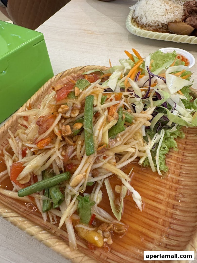 A close-up of a delicious Som Tum salad served in a woven basket, with a dish of rice and meat in the background.