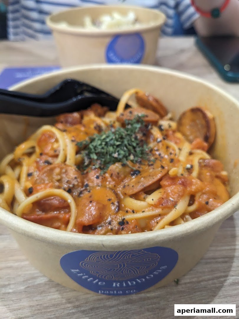 A close-up of a pasta dish with sausage and sauce in a takeaway bowl.