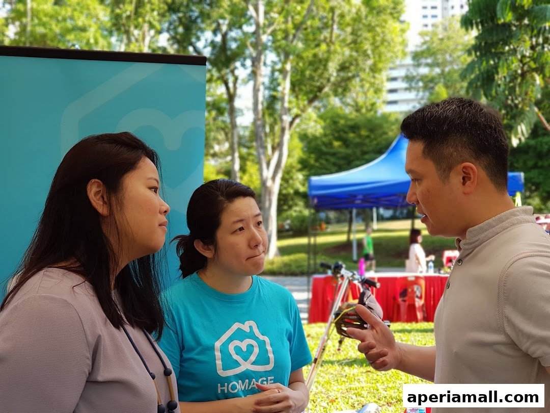 Three people are engaged in a conversation outdoors, with a blue tent and trees in the background.