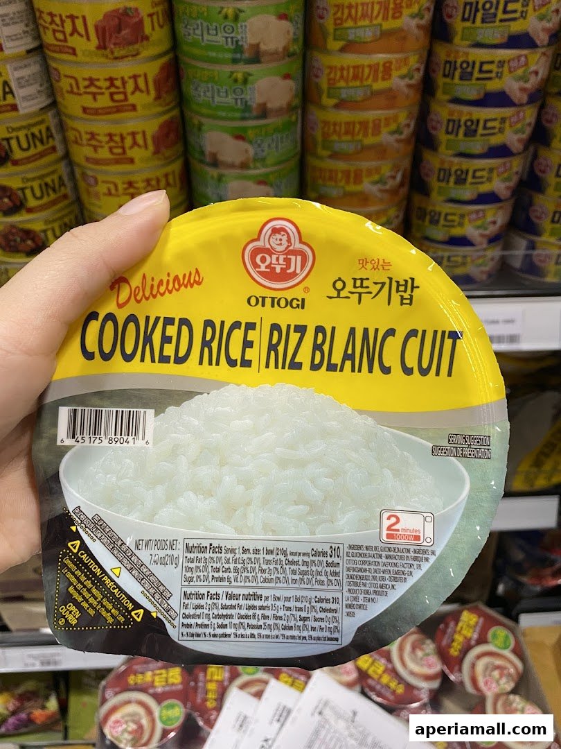 A hand holds a package of Ottogi Cooked Rice with other Korean food products in the background.