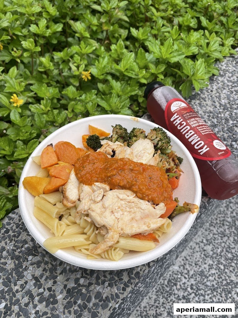 A healthy meal bowl with pasta, chicken, vegetables, and a bottle of kombucha.