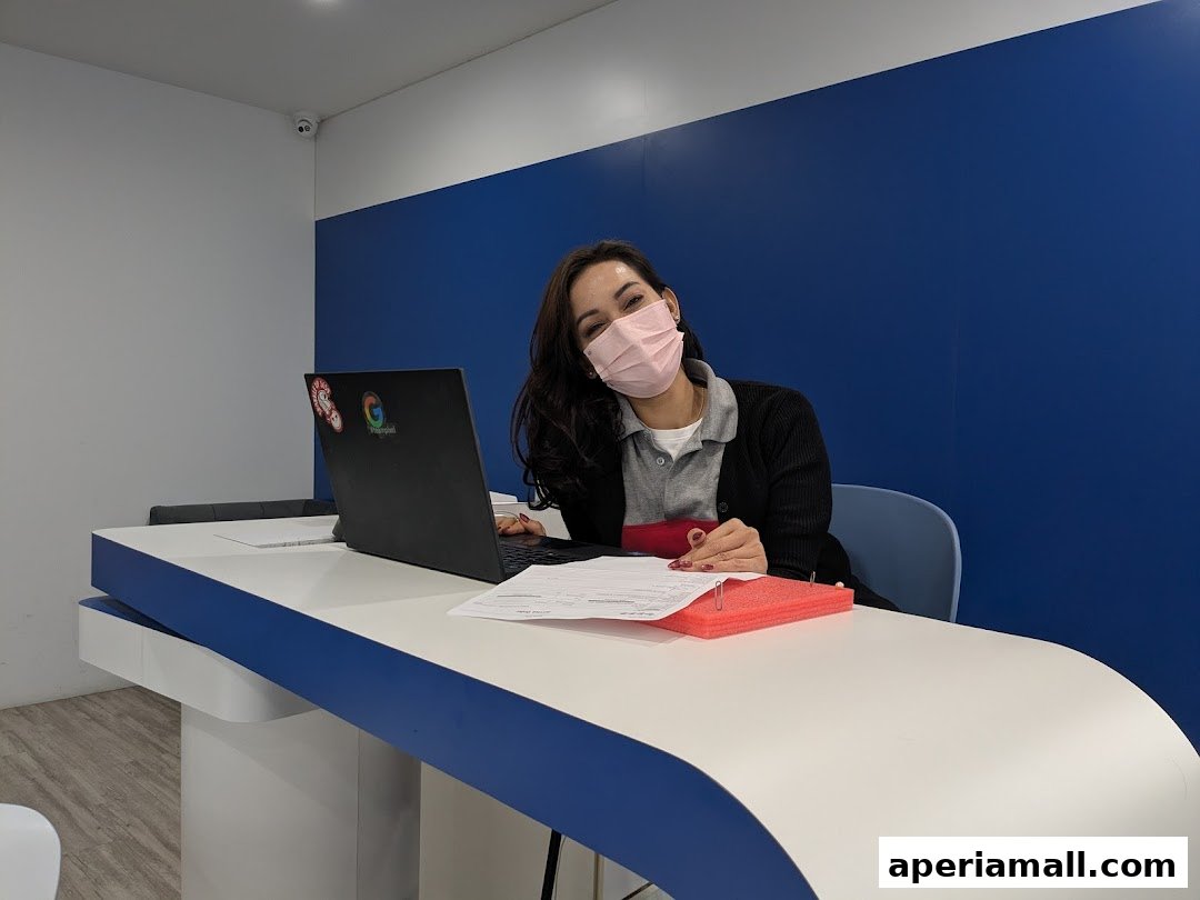 A woman wearing a mask sits at a desk with a laptop and papers.