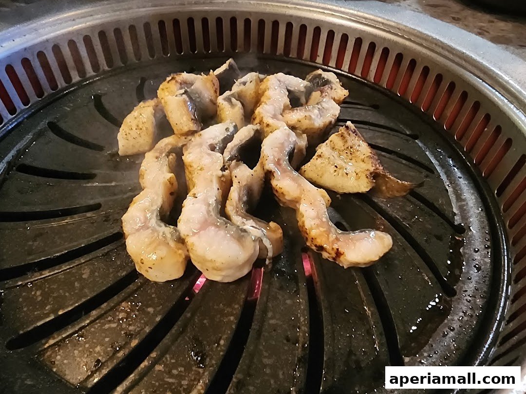 Sizzling pieces of seasoned eel are grilled on a round barbecue grill with visible heat.
