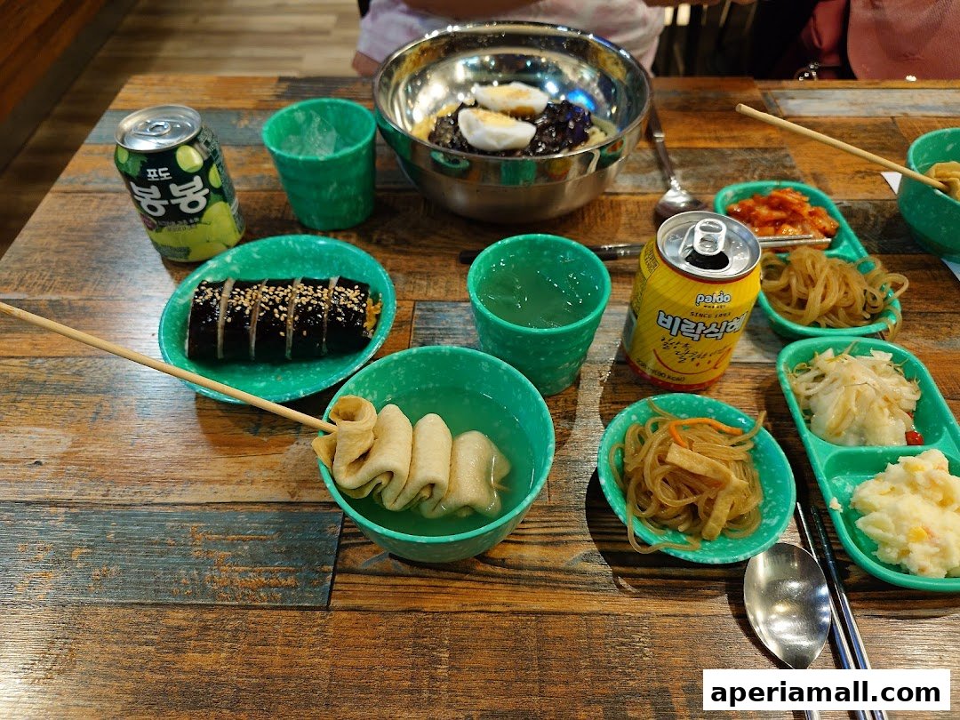 A table laden with Korean dishes, including kimbap, fish cakes, and various side dishes, with drinks.