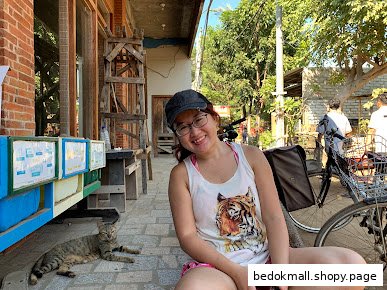 Smiling person and cat on pavement outside building with displays.