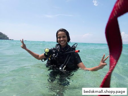 A smiling person in scuba gear stands in clear blue ocean water, giving peace signs.