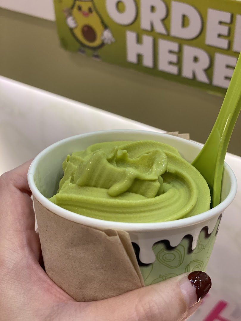 Hand holding a cup of green frozen dessert with a spoon, 'ORDER HERE' sign in background.