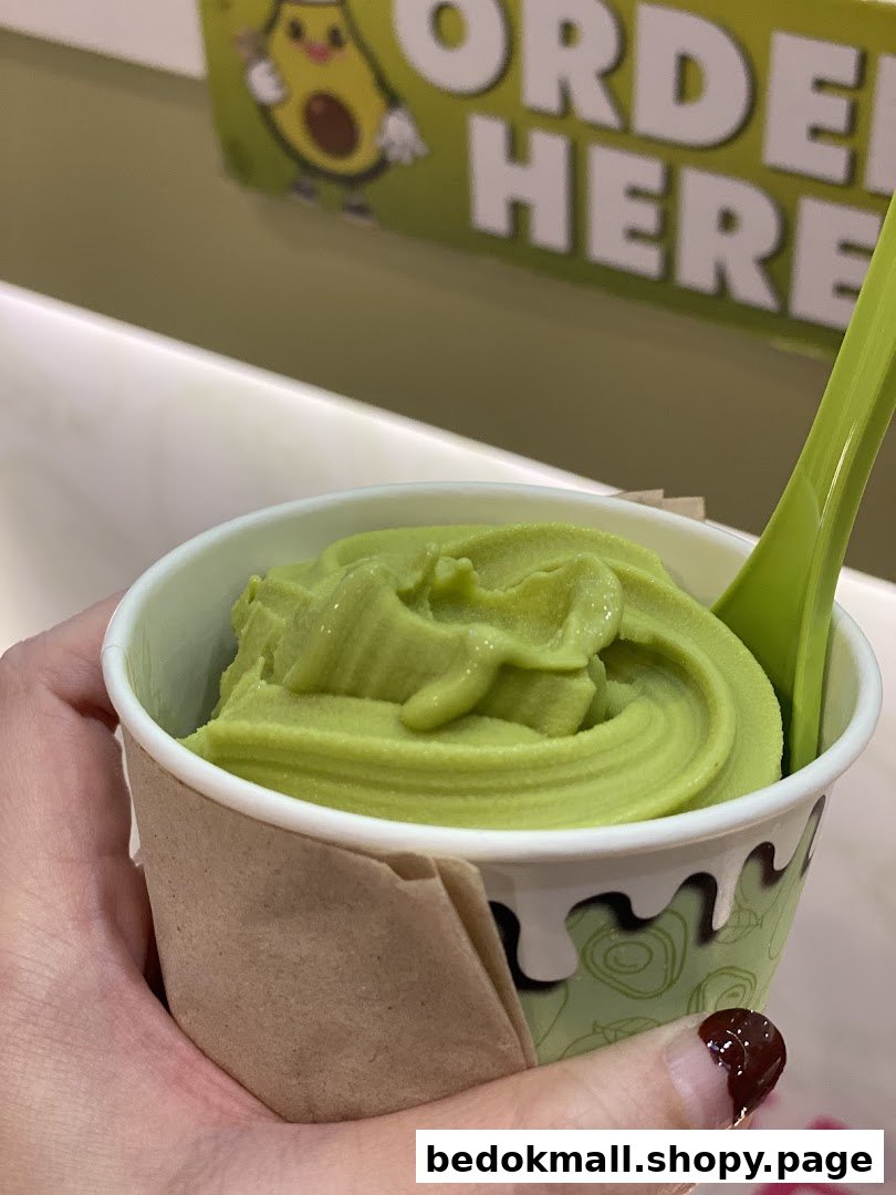 Hand holding a cup of green frozen dessert with a spoon, 'ORDER HERE' sign in background.