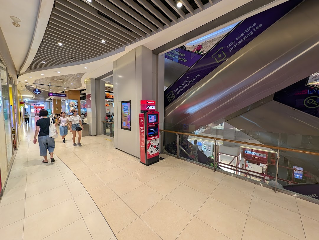 An AXS kiosk stands in a mall hallway with people and escalators nearby.