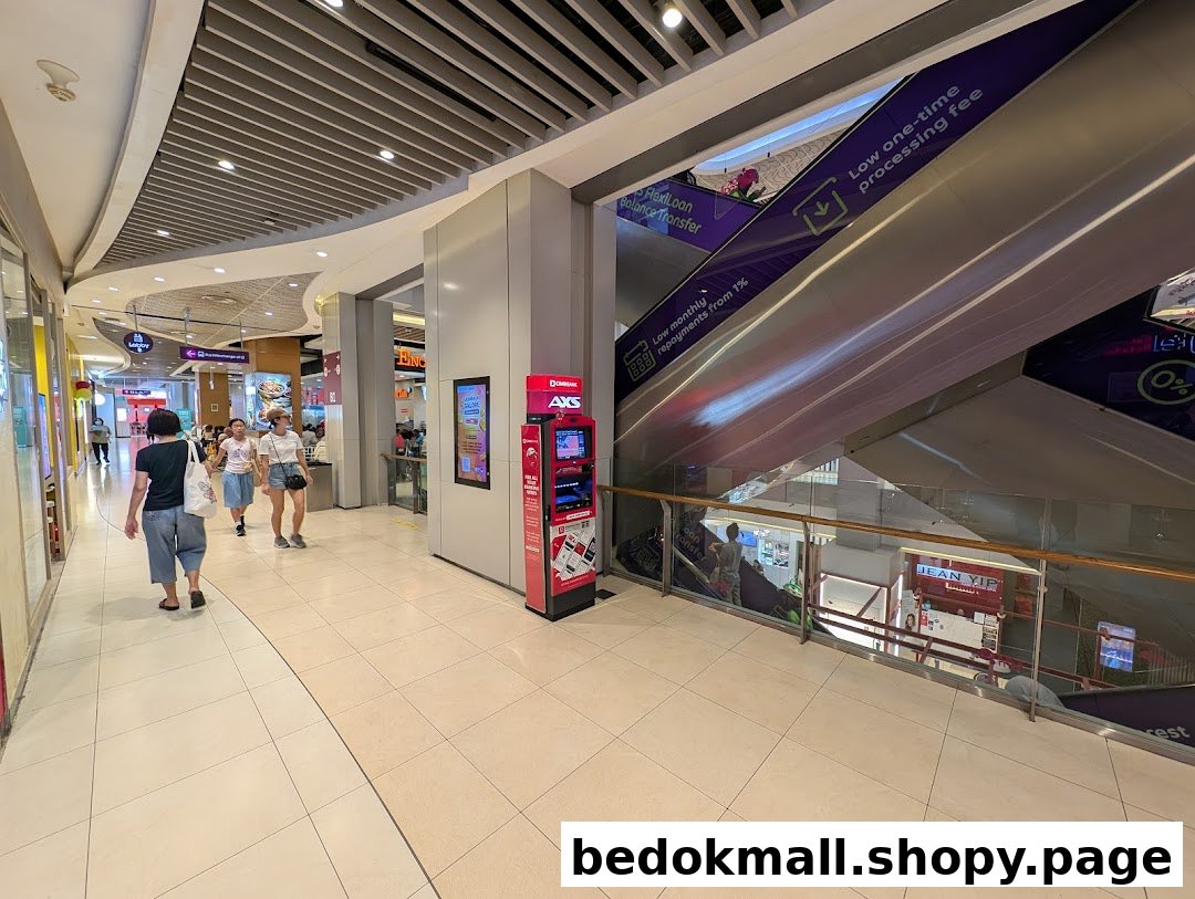An AXS kiosk stands in a mall hallway with people and escalators nearby.