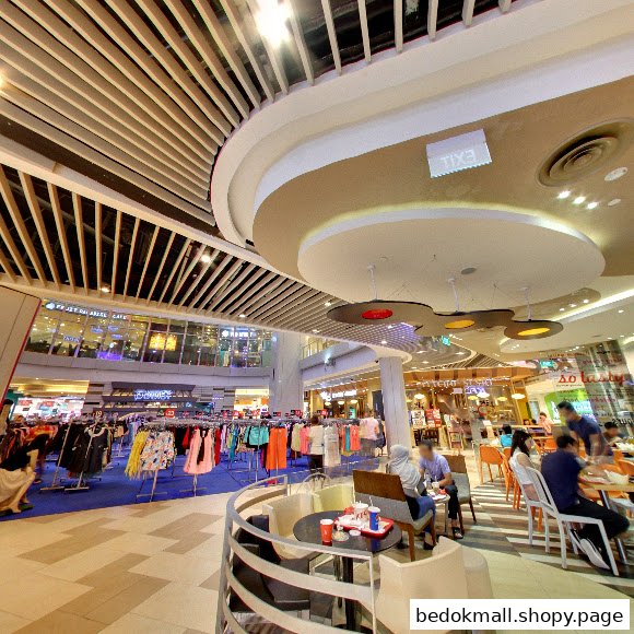 Interior of a shopping mall with clothing displays, a seating area, and people.