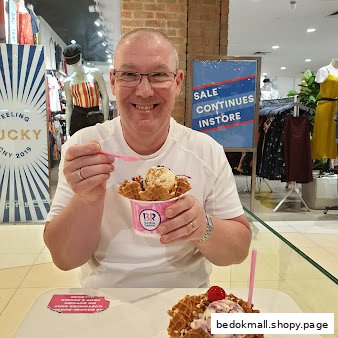 A man smiles while eating ice cream in waffle bowls inside a mall.
