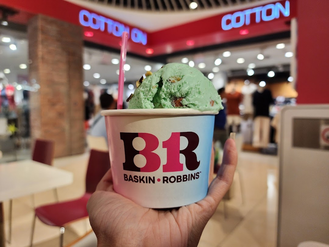 A hand holds a Baskin-Robbins ice cream cup with green ice cream in a mall.