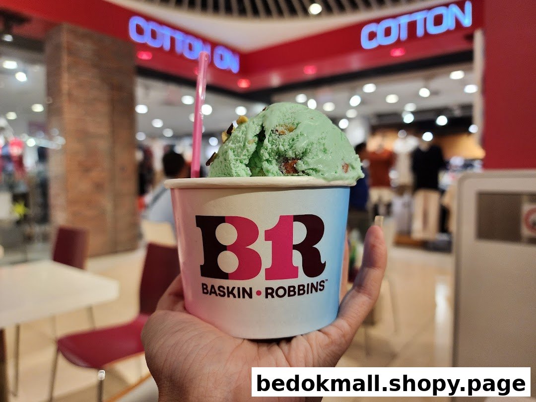 A hand holds a Baskin-Robbins ice cream cup with green ice cream in a mall.