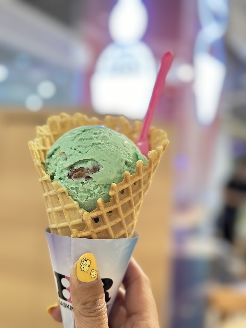 A hand holds a green ice cream scoop in a waffle cone with a pink spoon.