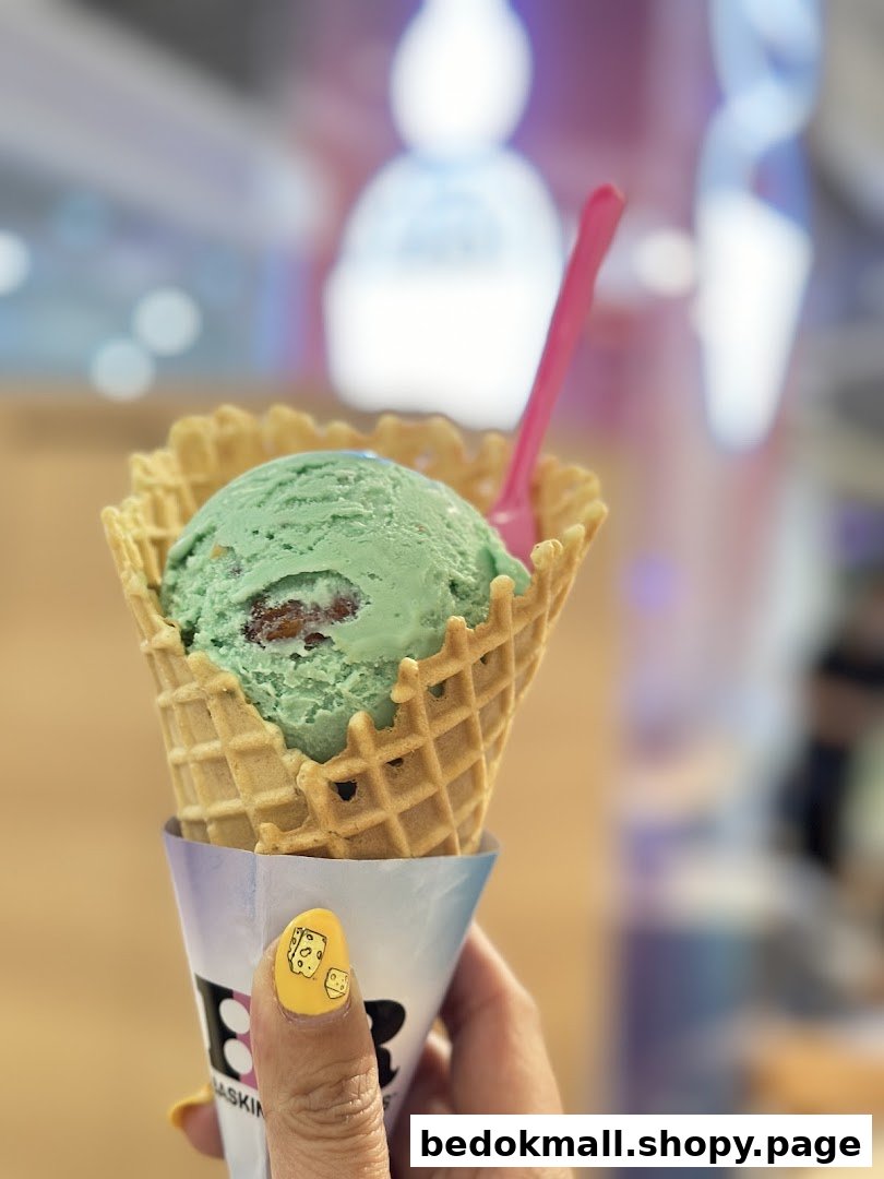 A hand holds a green ice cream scoop in a waffle cone with a pink spoon.