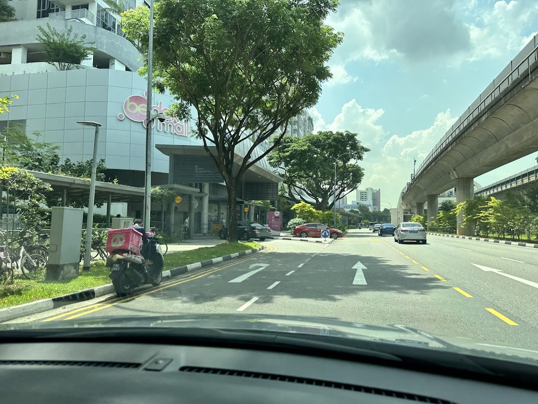 Exterior view of Bedok Mall from a street with cars, a scooter, and elevated tracks.