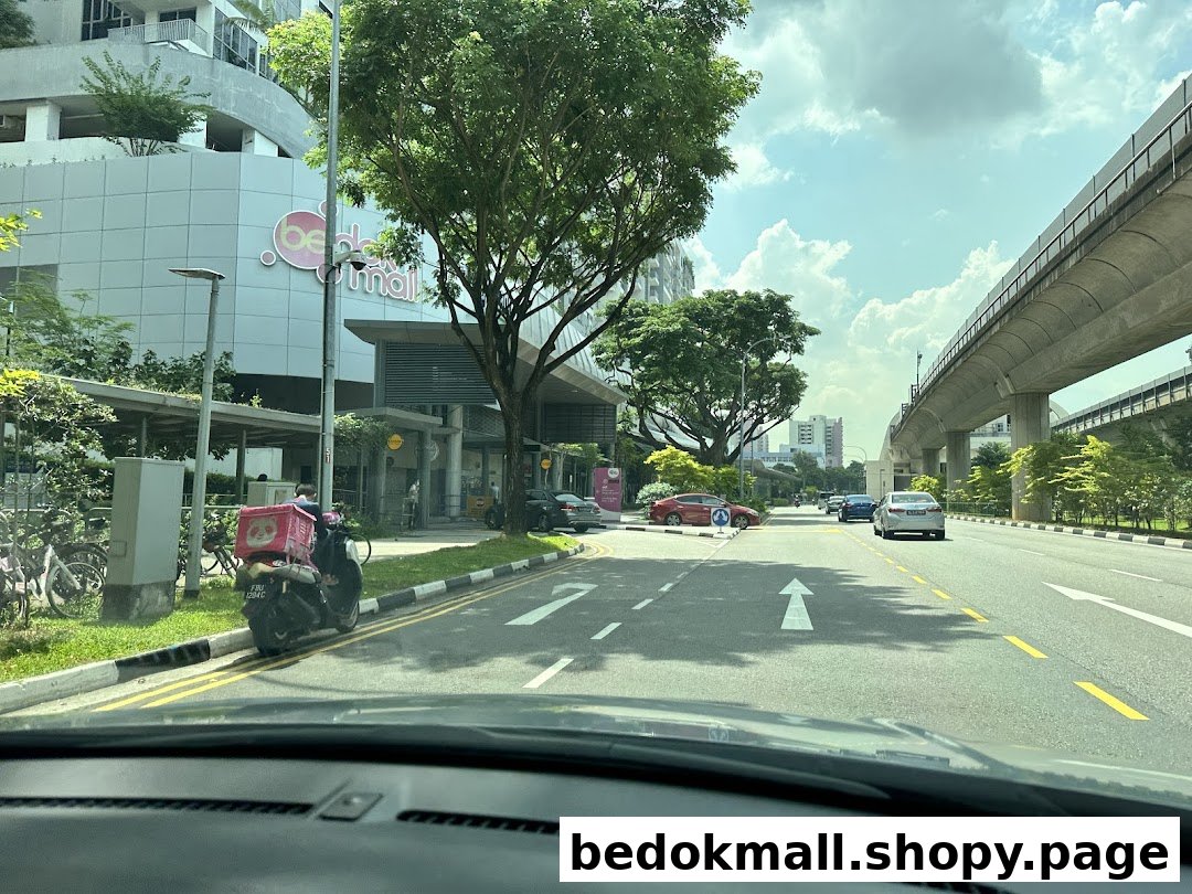 Exterior view of Bedok Mall from a street with cars, a scooter, and elevated tracks.