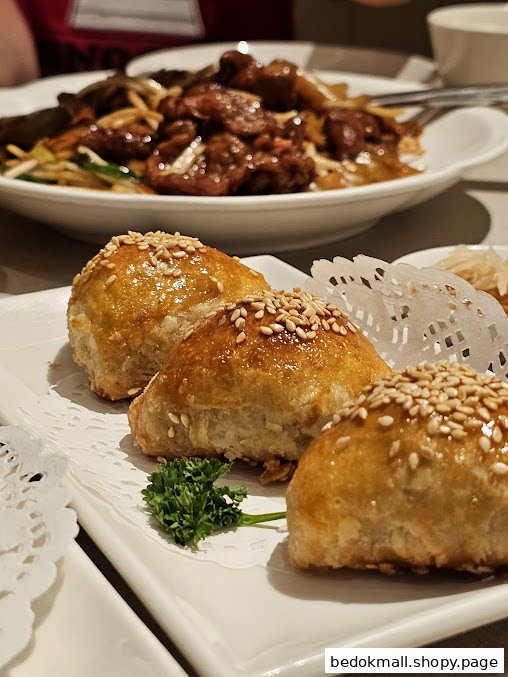 Close-up of three golden pastries with sesame seeds, with a stir-fried dish in the background.