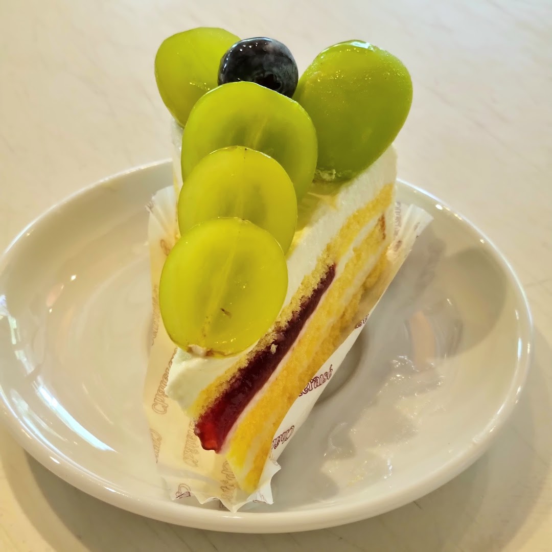 A slice of cake topped with green and one dark grape on a white plate.