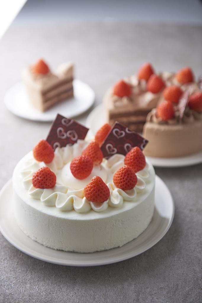 A white cream cake with strawberries and chocolate decorations, with other cakes in the background.