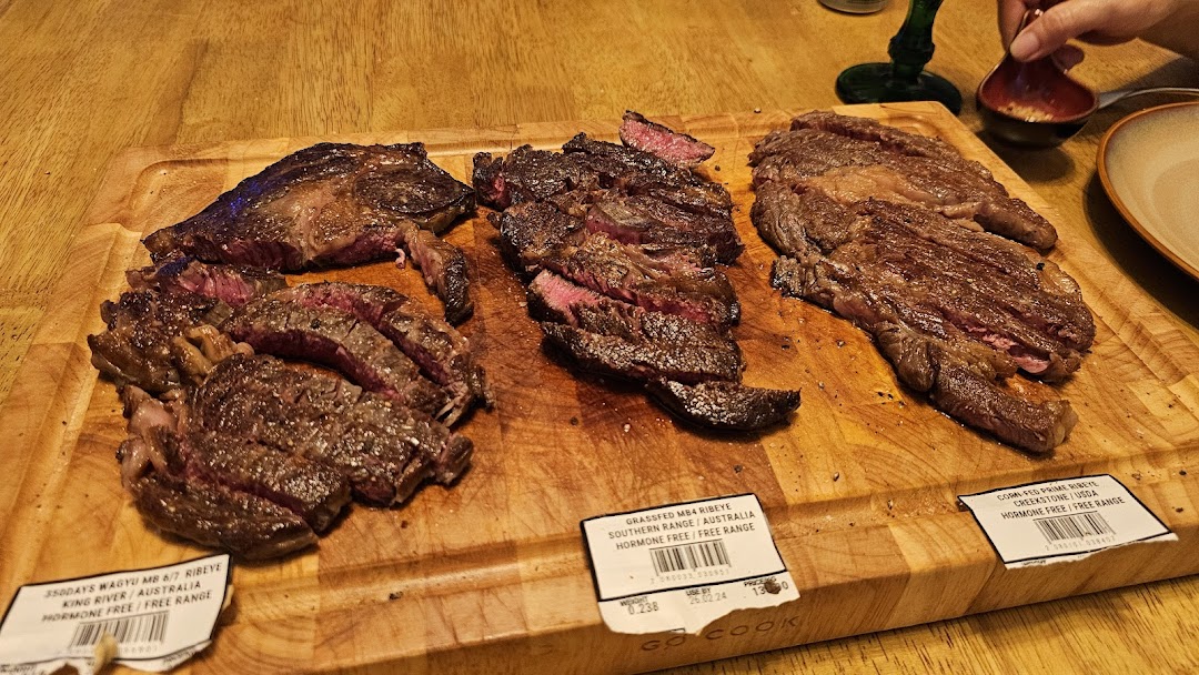 Three cooked steaks on a wooden board with descriptive labels.