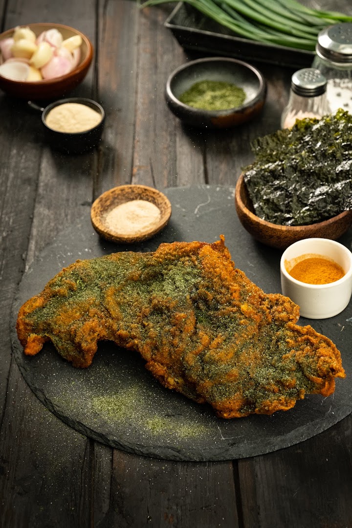 Golden fried chicken with green seasoning on a dark surface, surrounded by ingredients.