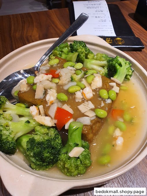 A dish of tofu, broccoli, and seafood in sauce with a spoon.