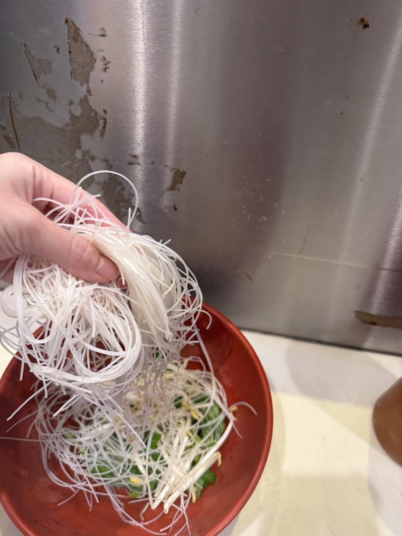 Hand adding white noodles to a red bowl with bean sprouts.