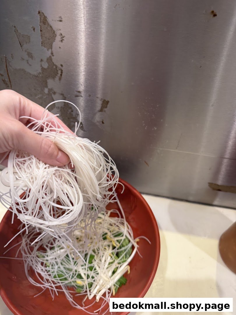 Hand adding white noodles to a red bowl with bean sprouts.