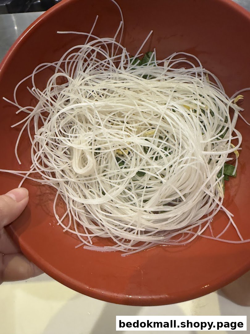 A red bowl filled with white vermicelli noodles and some greens.