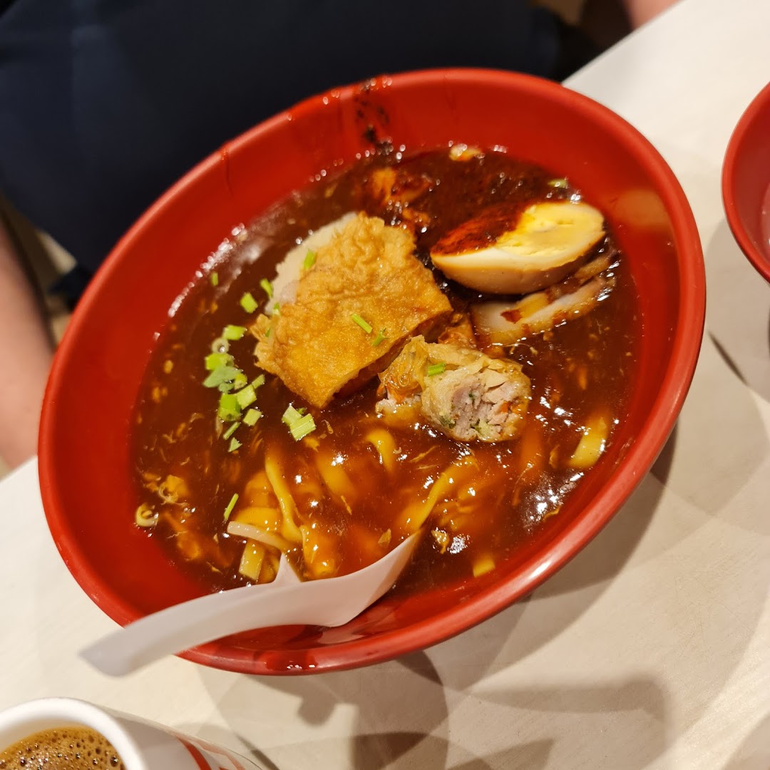 A red bowl of noodles with dark sauce, egg, and fried toppings.