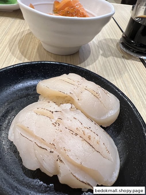 Two scallop sushi pieces on a black plate with salmon bowl in background.