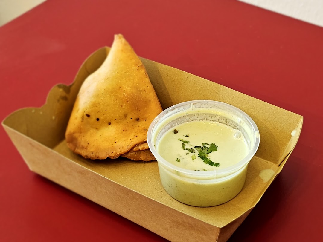 A samosa and a cup of green chutney in a cardboard tray on a red surface.