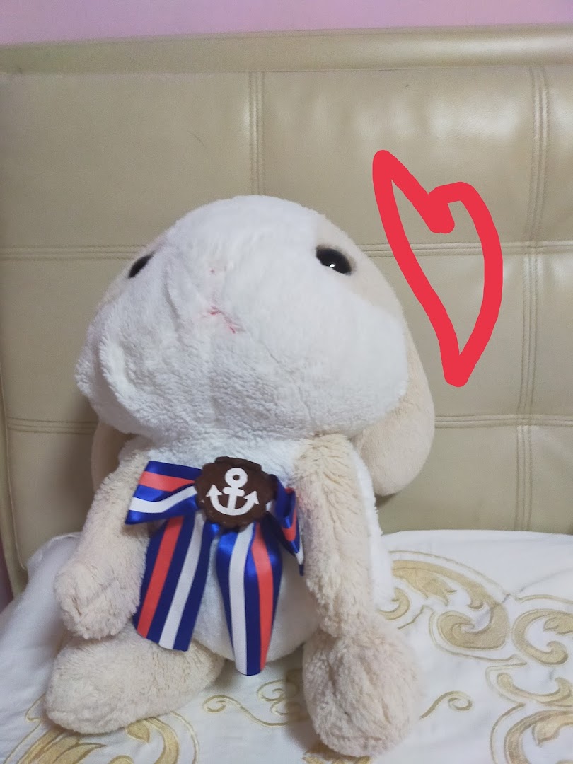 A light-colored plush toy with a nautical bow and anchor sits on a bed.