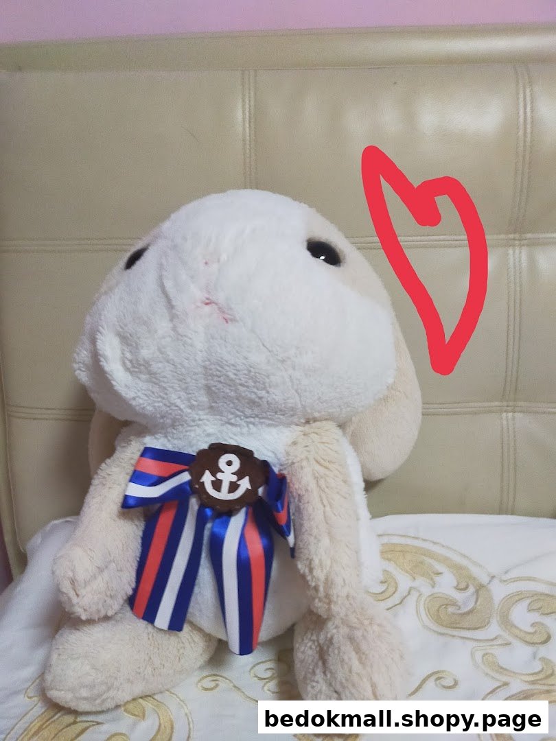 A light-colored plush toy with a nautical bow and anchor sits on a bed.