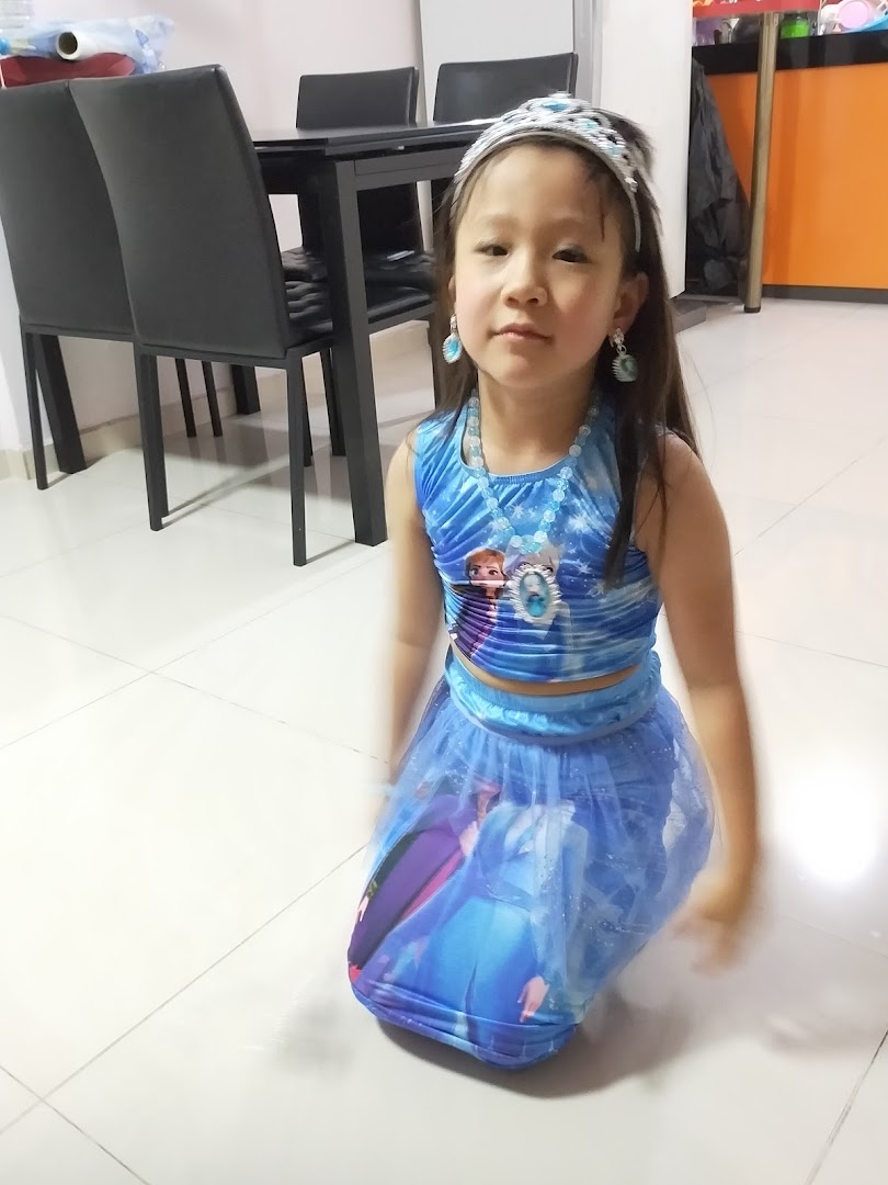 A young girl in a blue costume and tiara kneels on a tiled floor indoors.