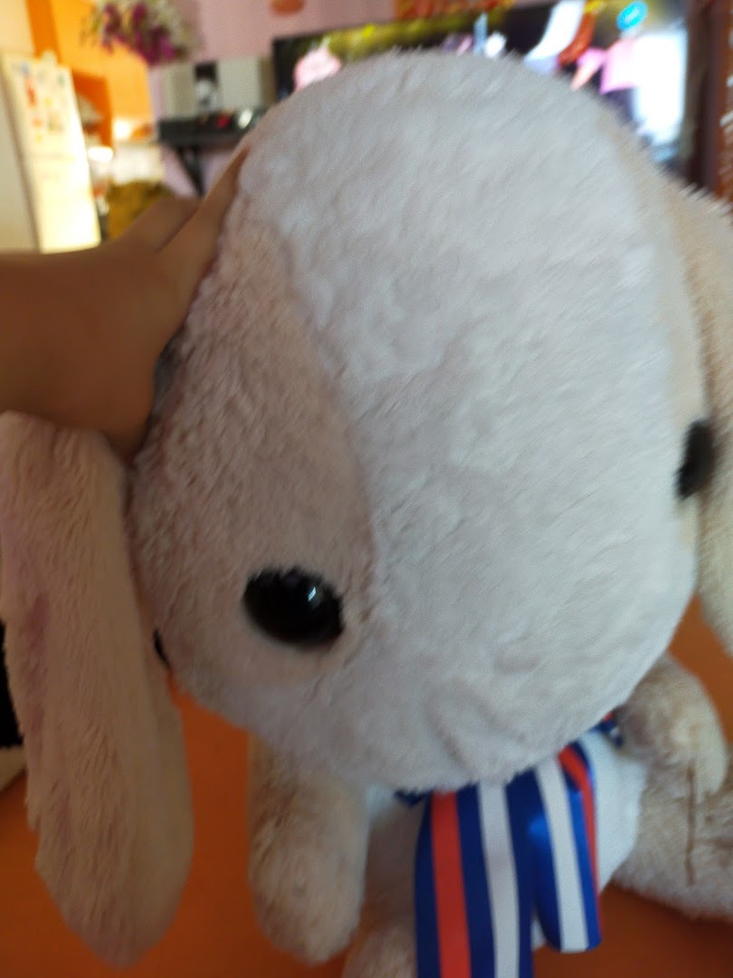A hand touching a large, light-colored plush toy with a striped ribbon.