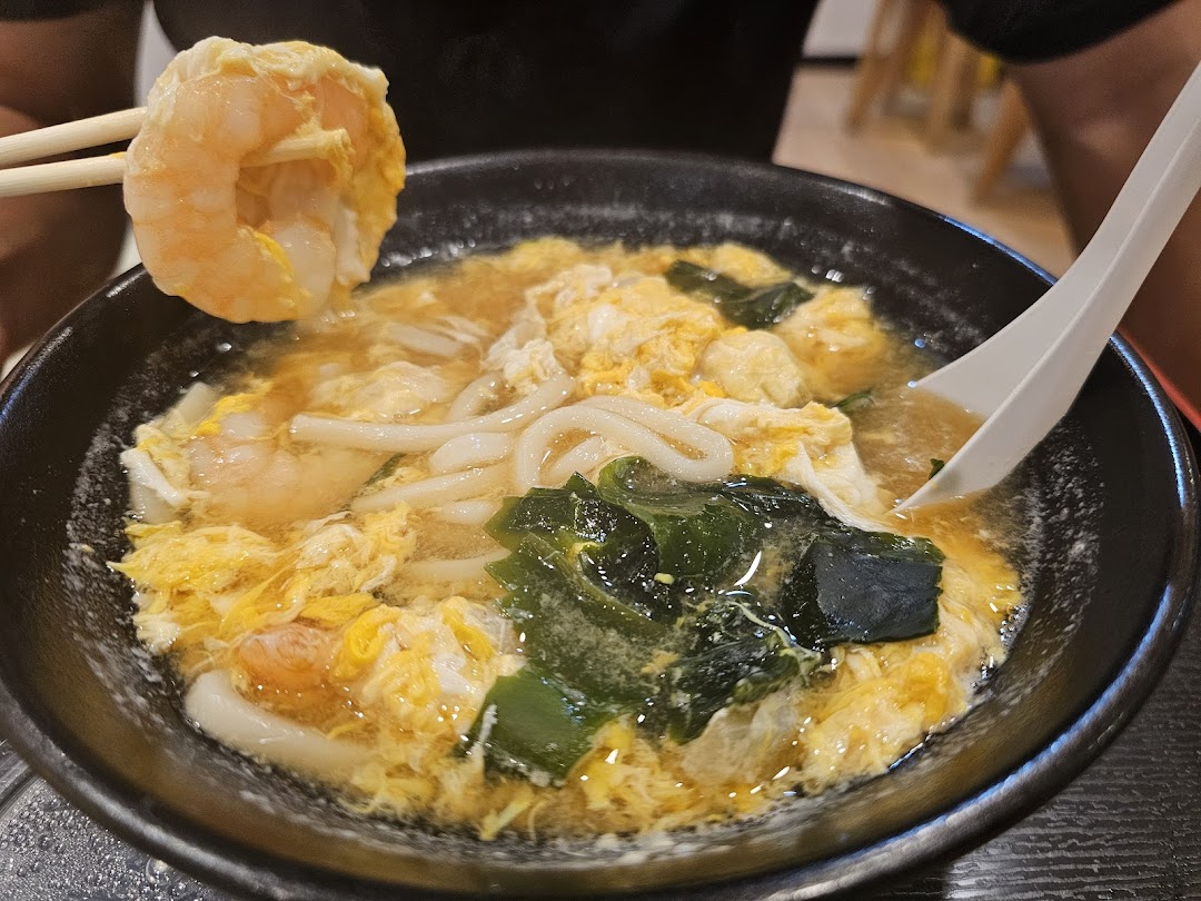 A close-up of a bowl of udon noodles with shrimp, egg, and seaweed.