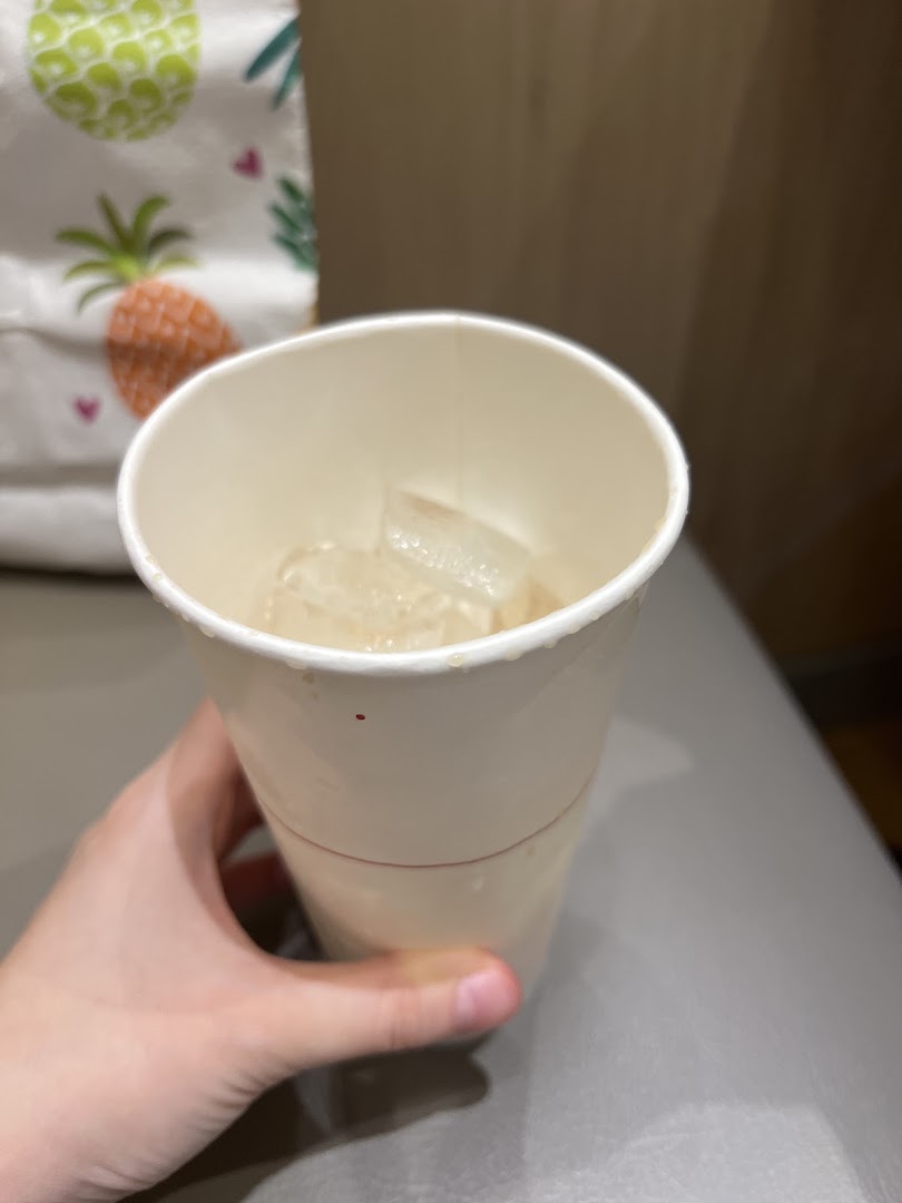 Hand holding a paper cup with ice, with a pineapple-patterned bag in background.